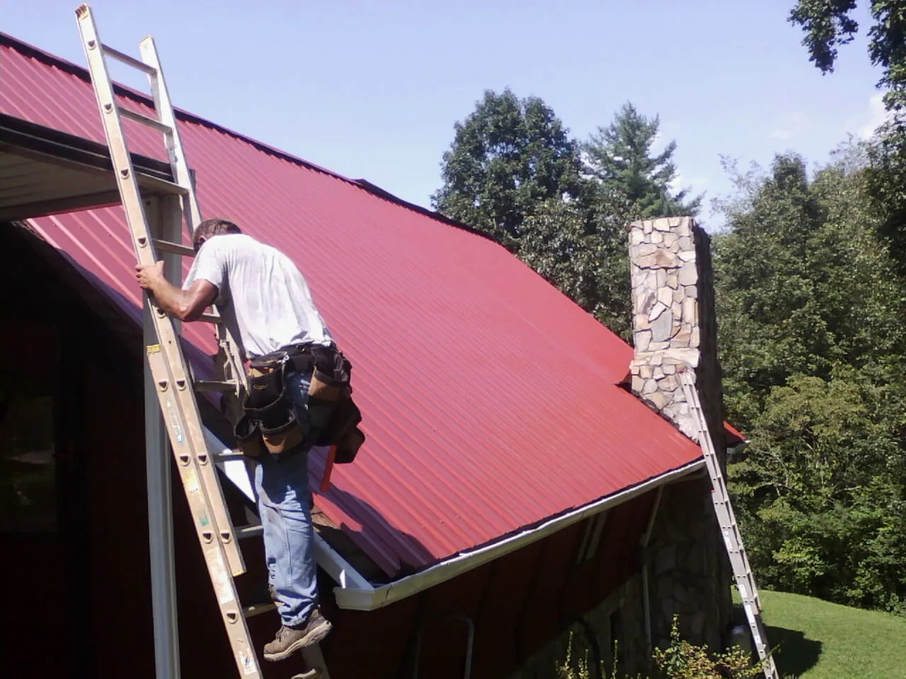 Roofer on a ladder beside a red metal roof and stone chimney — Soffit & Fascia Repair by Roofing Brothers in Fruitland